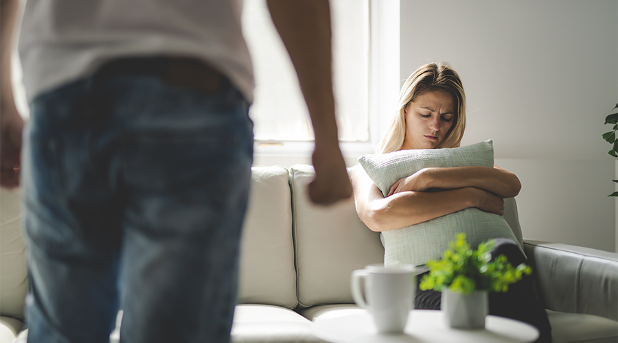 A woman sits on a sofa holding a pillow, with part of a person standing in the foreground.