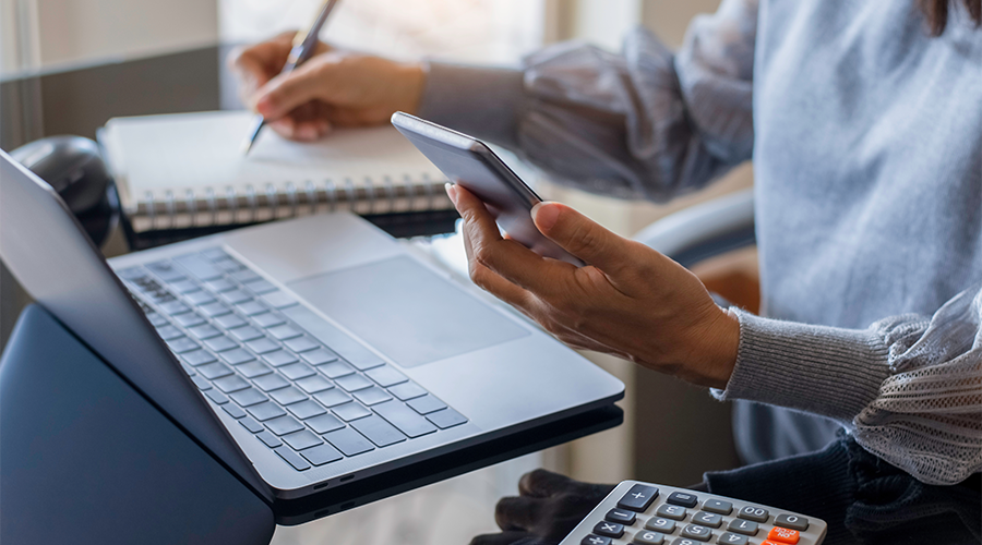 Person multitasking at a desk with a laptop, smartphone, notebook, and calculator.
