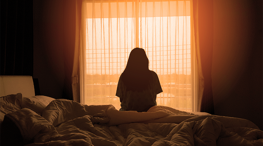 Silhouette of a person sitting on a bed in front of a sunlit window with sheer curtains.