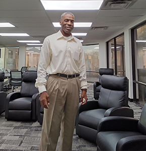 A man in a light dress shirt and beige trousers stands near black leather armchairs in a well-lit office space.