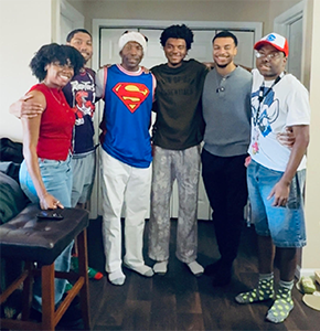 A group of six people standing inside a room, smiling at the camera, wearing casual outfits.