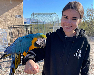 A young woman, with a colorful parrot perched on her arm, smiles for the camera.