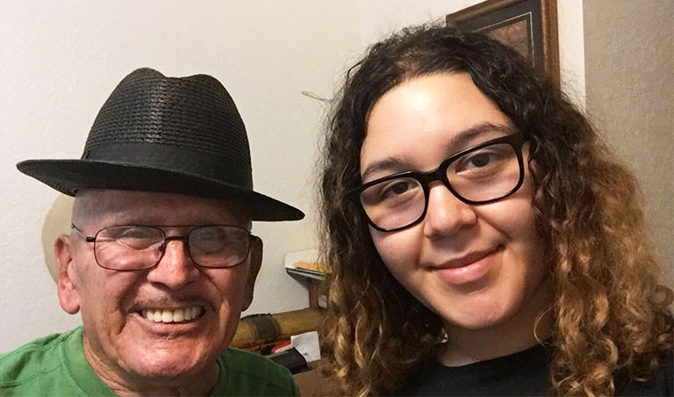 An elderly man wearing a hat and glasses stands next to a young girl wearing glasses. Both are smiling.