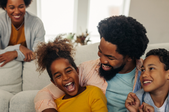 Father cuddles playfully with two young children as mother watches nearby.