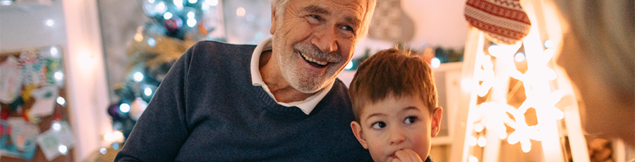 An older man in a sweater smiles next to a young child, in front of Christmas lights and decorations.