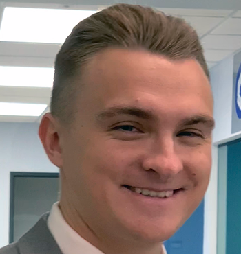 Man in a grey suit with a blue tie smiling indoors.