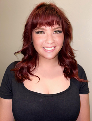 A person with auburn hair and a black shirt smiles at the camera against a plain, light-colored background. A person with auburn hair and a black shirt smiles at the camera against a plain, light-colored background.