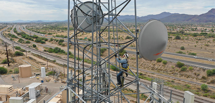 A worker climbs a telecommunications tower in a desert landscape with highways and distant mountains.