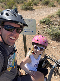 Adult and child in helmets smiling on a desert bike trail.