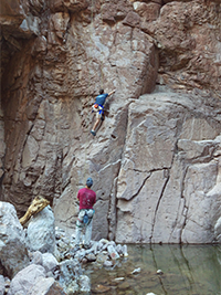 Person rock climbing on a vertical rock face with another person observing from below.