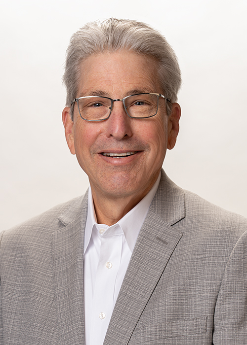 Man with gray hair and glasses wearing a gray blazer and white shirt against an off-white background.