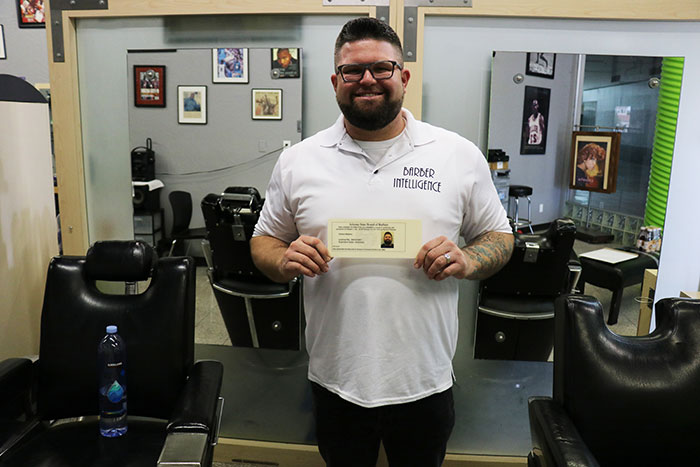 a man stands inside a barbershop; he is holding a certificate and smiling