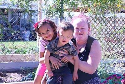 a little girl, toddler brother, and their mom pose in front of their vegetable garden.