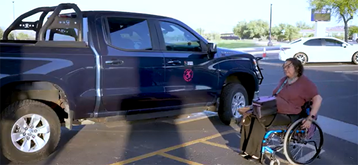 A person in a wheelchair next to a blue pickup truck in an accessible parking space.
