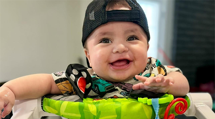 Smiling baby in a colorful jumper seat wearing a backwards black cap.