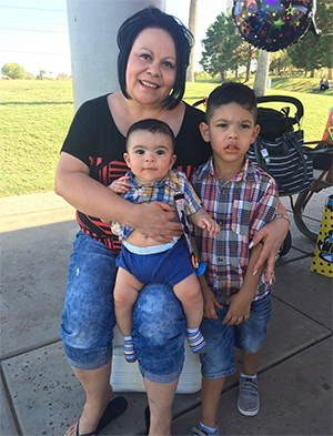 A woman with two children at an outdoor park, one seated on her lap and another standing beside her.