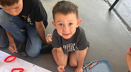 A young child sits on a concrete floor with crayons and paper, looking at the camera.