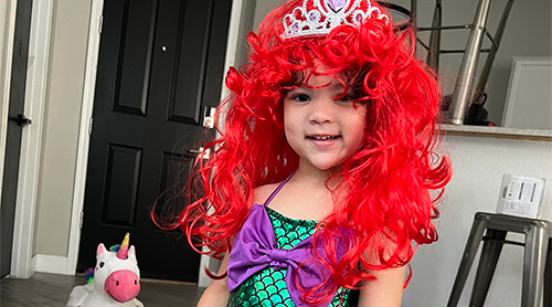 Child in a bright red wig, mermaid costume, and tiara, with a toy unicorn in the background.