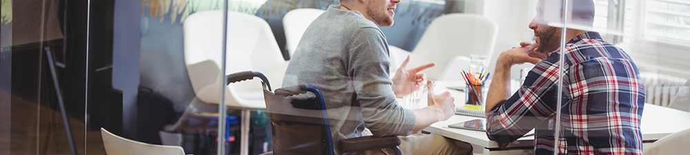 Disabled man in wheelchair at work talking to another male coworker at a table.