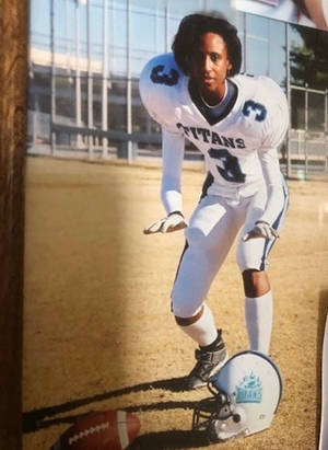 A young person in a white football uniform crouches on a field with a helmet and football nearby.