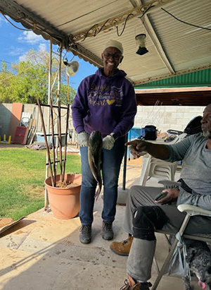 Person standing on a porch holding two large fish, with fishing rods in the background and another person seated nearby.