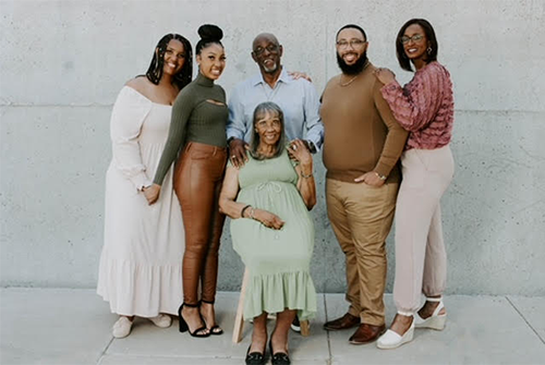 A group of six people, five standing and one seated, smiling together in front of a concrete wall.