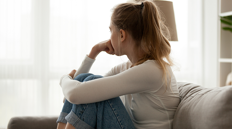 Side view young woman looking away at window sitting on couch at home.