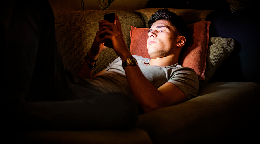 Young Man Lying on Sofa at Night and Illuminated by Light from Screen of Cell Phone at Home in Living Room