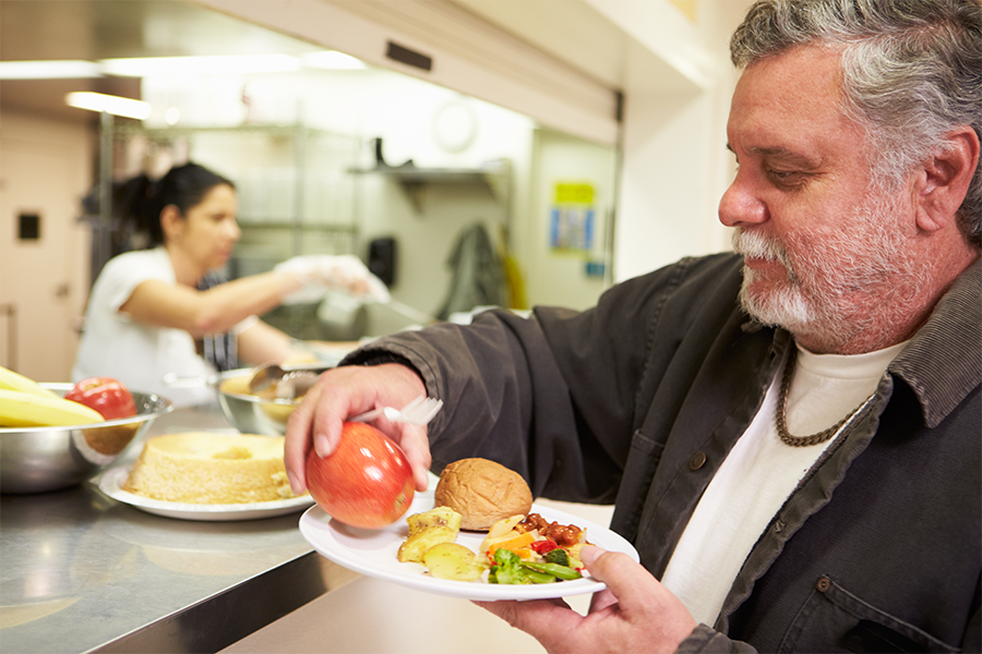 Man selecting food at a cafeteria serving counter, holding a plate with an apple and other foods.