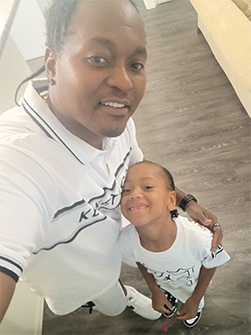 Person and child smiling for a selfie on a wooden floor, both in white clothes.