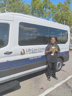 A man in a black outfit stands next to a white and blue van, smiling and giving thumbs up.