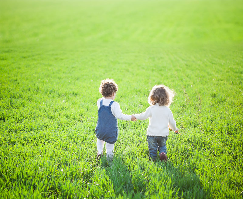 two toddler girls walk over a grassy field holding hands