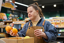 Young woman working in the produce section of a grocery store holding two oranges.