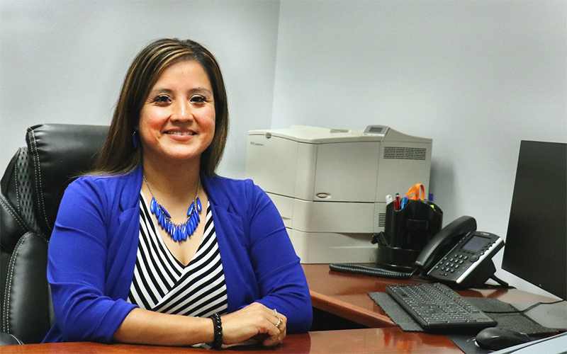 Woman in blue jacket sits at a desk in a workplace setting.