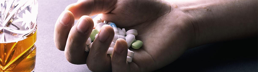 A hand holding various pills next to a glass of amber liquid on a dark surface.