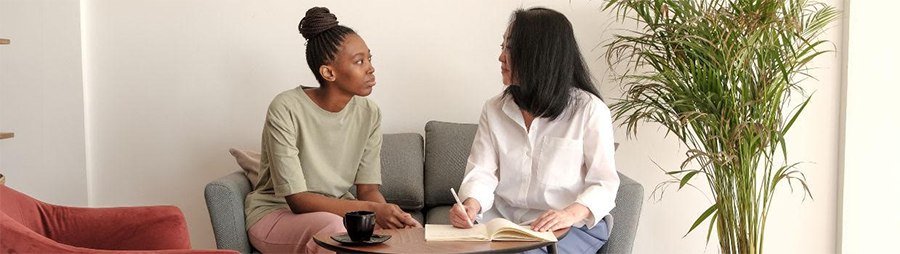 Two people sitting on a sofa, conversing with a notebook, a coffee table, and a plant nearby.
