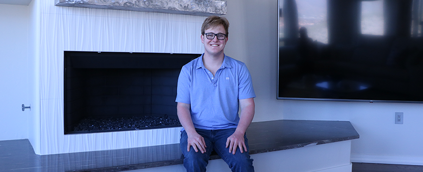 young man dressed in jeans and a polo shirt, sits on the ledge of his living room fireplace