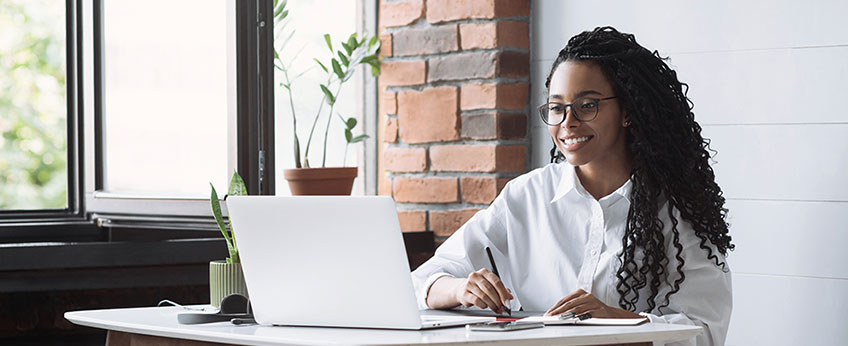 young woman sitting at a desk, writing on a notebook; in front of her, a laptop computer is open