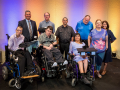 Six men and three women pose together for a picture. There are three individuals using wheel chairs in the front row, while the other participants stand in a row behind them.
