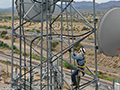 A worker climbs a telecommunications tower with antennas, set against a landscape of highways and mountains.