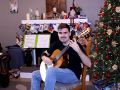 Young man, sitting in front of a Christmas tree, plays an acoustic guitar