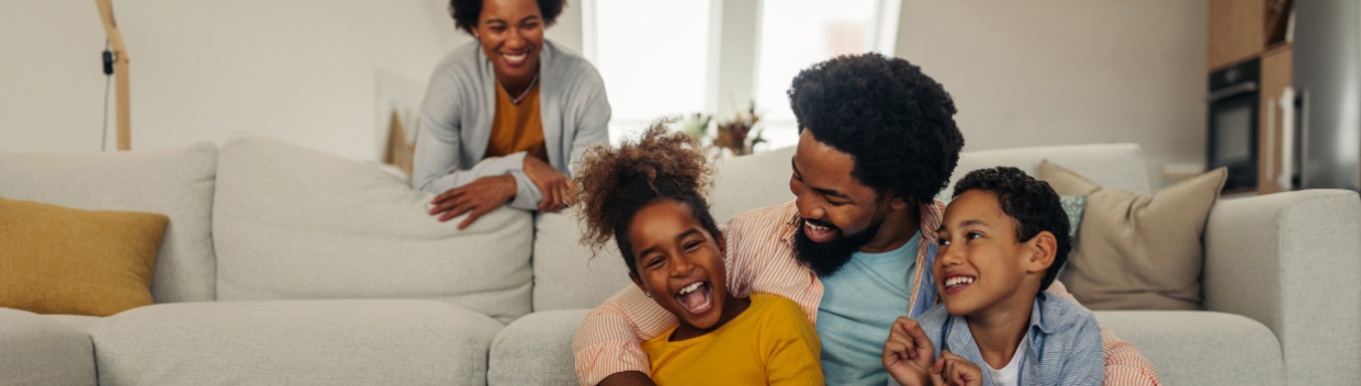Father cuddles playfully with two young children as mother watches nearby.