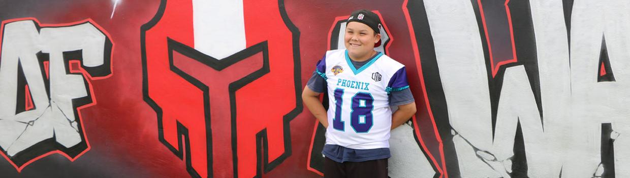 Boy wearing his team’s jersey stands in front of a painted wall.