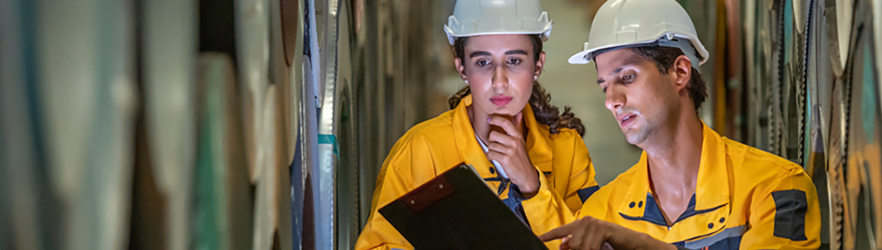 Two workers in safety helmets and industrial uniforms review a clipboard.