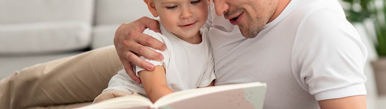 A smiling man reads a book to a smiling child as they are relaxing.