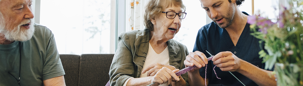 Two older adults teaching a young man how to knit