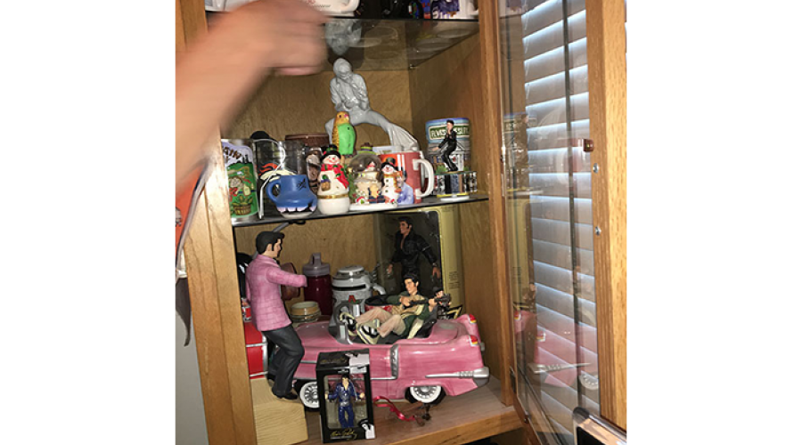 a  hand is reaching into a curio cabinet; the shelves are filled with cups and Elvis memorabilia