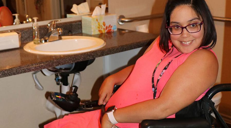 A woman sits in a while chair, smiling, in front of an accessible bathroom sink.