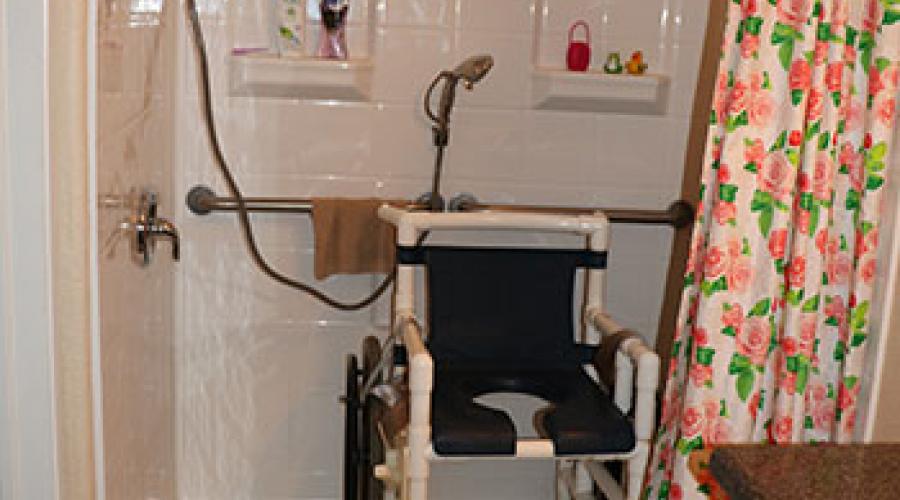 A roll-in shower with a wheelchair inside of it. A handle runs along the back. The shower has a hand faucet.