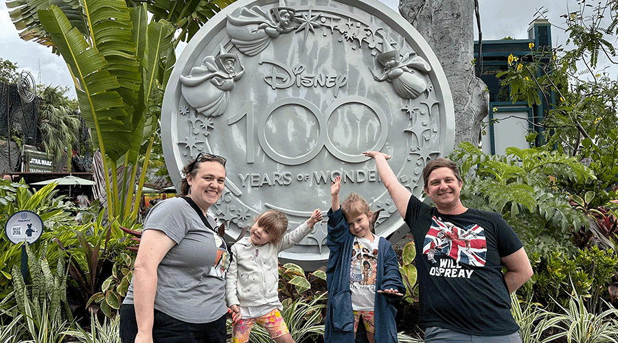 woman, man and two small children standing before a large coin that reads "Disney 100 Years of Wonder"